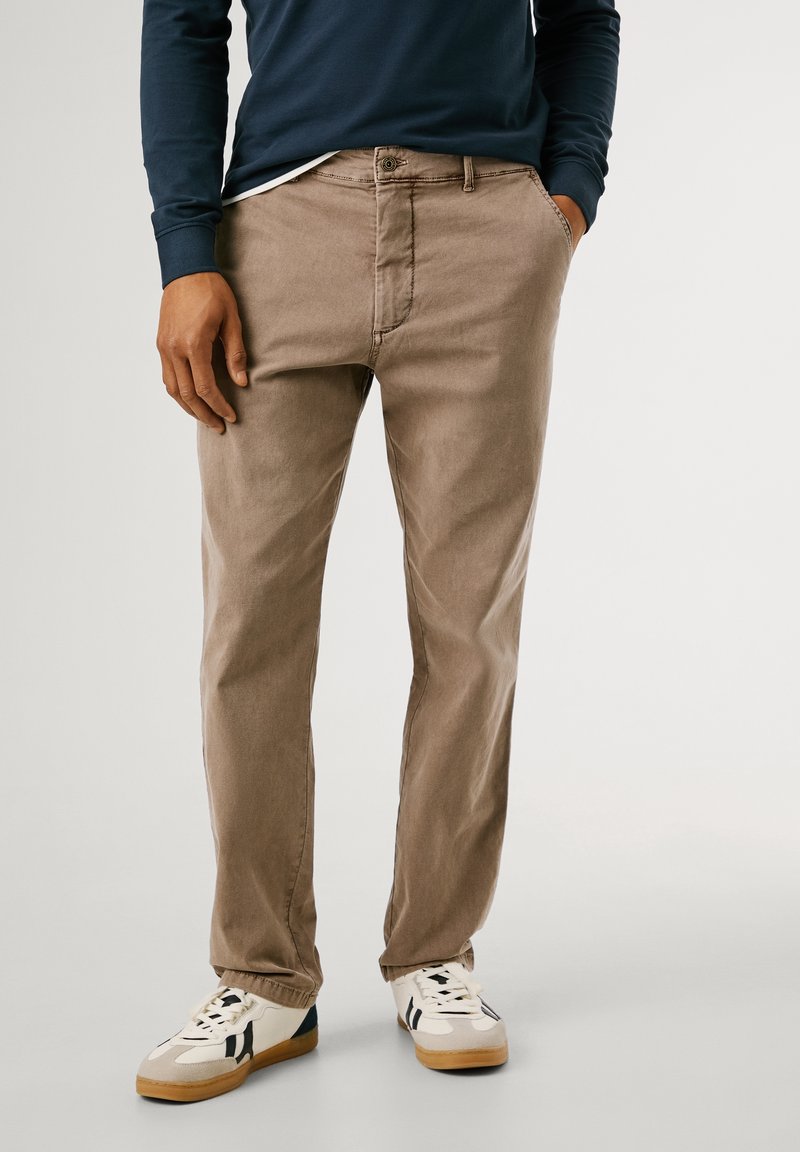 Homme portant un pantalon beige coupe décontractée, des baskets blanches avec des détails noirs, et une chemise manches longues bleu marine, debout les mains dans les poches.