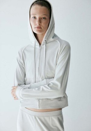 Joven mujer de piel clara y ojos azules lleva una sudadera blanca con cremallera y capucha puesta, junto con pantalones a juego sobre un fondo liso.