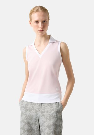 Jeune femme portant un haut sans manches col chemise rose pâle et un pantalon gris avec un léger motif de palmiers, debout les mains dans les poches.