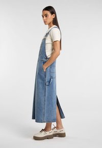 Vestido de tirantes de mezclilla, azul claro, hasta la rodilla con aberturas laterales, bolsillos frontales, corsé ajustado, tirantes ajustables, combinado con zapatos gruesos en color beige.