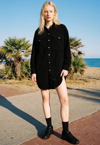 Blonde Frau steht auf einem gepflasterten Weg nahe dem Strand, trägt ein schwarzes Hemdkleid mit Knöpfen, schwarze Socken und schwarze Sneaker unter klarem blauem Himmel.