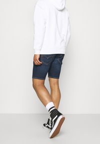 Tummansinisiä farkkushortseja suoran leikkauksen ja näkyvien ompelujen kanssa. Yhdistetty valkoisen hupparin ja mustien korkeavarrellisten tennareiden kanssa.