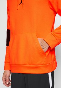 Sudadera naranja con un acento negro en la manga, bolsillo tipo canguro y cuerdas para la capucha. Textura suave y corte atlético.