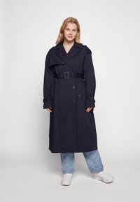 Trench coat blu navy con design doppiopetto, ampio colletto, cintura anteriore e polsini regolabili. Tessuto liscio con dettagli minimi.