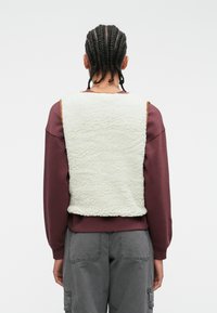 Fleece vest in off-white met een textured oppervlak, gedragen over een bordeauxkleurig langemouwen shirt, gecombineerd met grijze cargobroek.