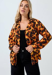 Camicia a maniche lunghe in tessuto nero e arancione a motivi, con rever a punta e tasca frontale, abbinata a pantaloni neri a vita alta.