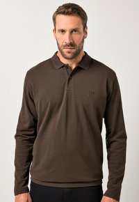 Camisa polo marrón de manga larga con textura acanalada, una tapeta de dos botones y un logo sutil en el pecho, combinada con jeans oscuros.