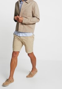 Giacca di cotone beige sopra una camicia a quadri blu, abbinata a pantaloni corti beige chiaro e scarpe da barca color nocciola. Outfit casual da esterno, con tasche.