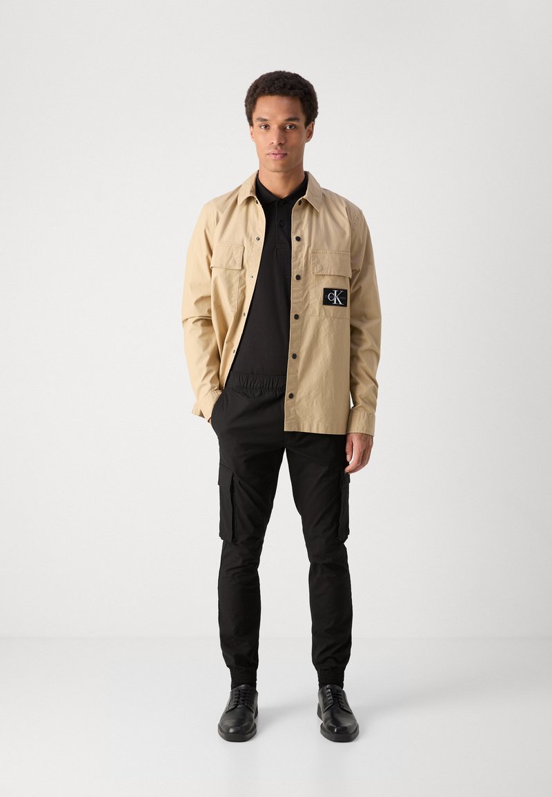 Camicia beige con bottoni e due tasche frontali sopra una polo nera, abbinata a pantaloni cargo neri e scarpe nere. Tessuto morbido, vestibilità casual.