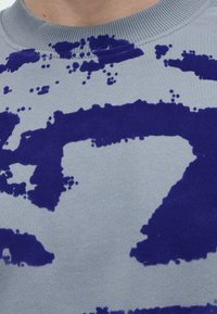 Gros plan d'un pull gris clair avec des motifs abstraits bleu foncé irréguliers, porté par une personne.