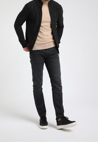 Czarna kurtka na zamek z paskami na beżowym, teksturowanym swetrze, w połączeniu z czarnymi dżinsami i czarnymi trampkami z białymi podeszwami, na neutralnym tle.