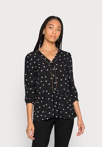 Blusa preta com bainha peplum, apresentando bolinhas coloridas, decote em V e fecho com botões à frente. Mangas longas com punhos elásticos.