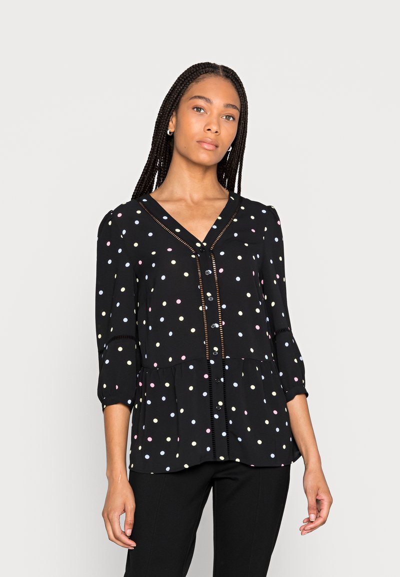 Blusa preta com bainha peplum, apresentando bolinhas coloridas, decote em V e fecho com botões à frente. Mangas longas com punhos elásticos.