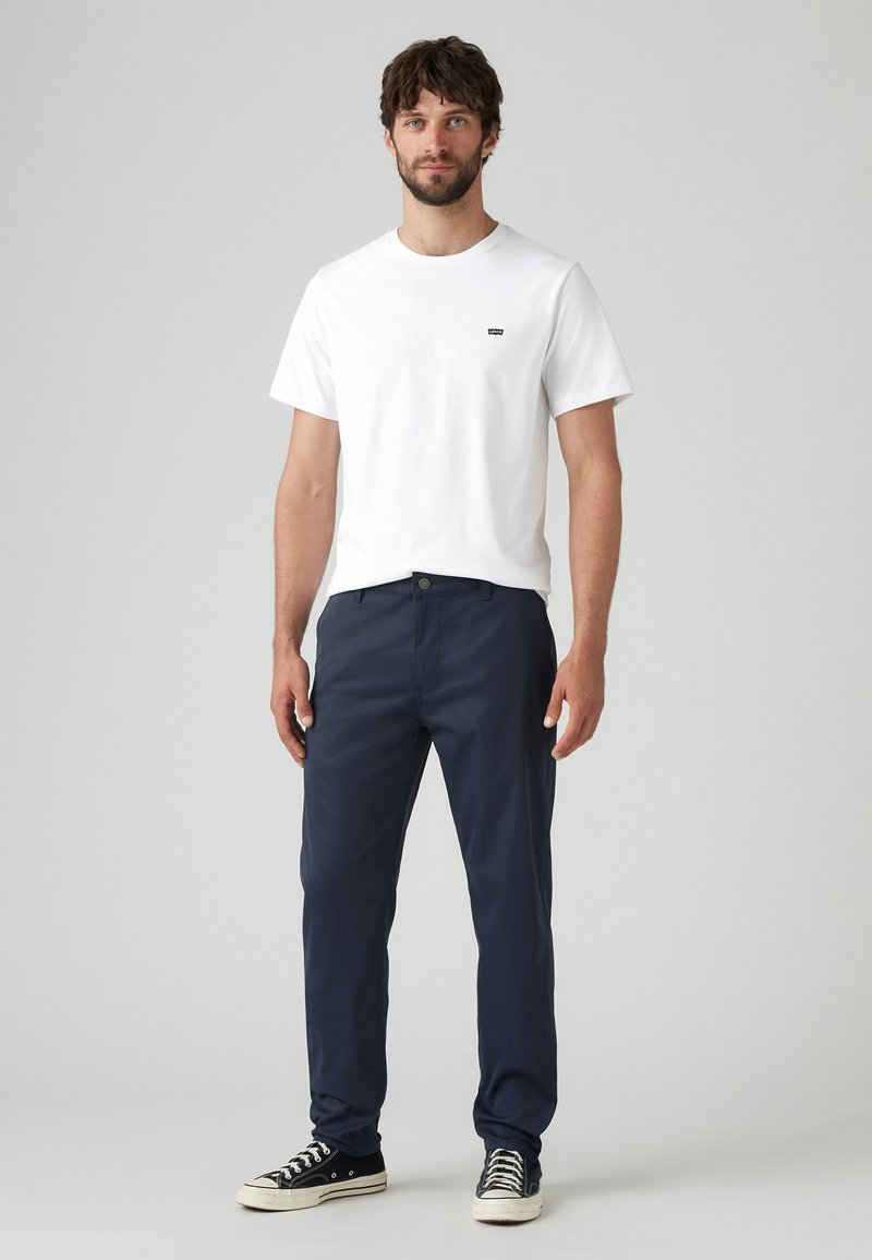 Tricou alb cu logo, pantaloni bleumarin și adidași negri. Croială slim, textură netedă, design basic fără modele sau accente.