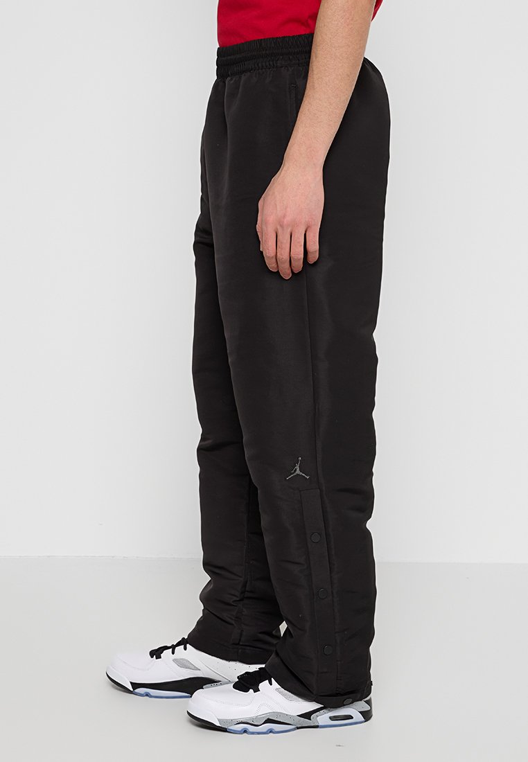 Schwarze Sporthose mit elastischem Bund, Seitenschnappverschlüssen und einem kleinen grauen Logo am Saum. Glatte Textur, lockere Passform.