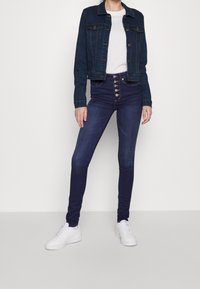Mörk denimjacka med knäppning, kombinerad med högmidjade mörkblå skinny jeans med femknappsframsida. Vita sneakers fulländar looken.