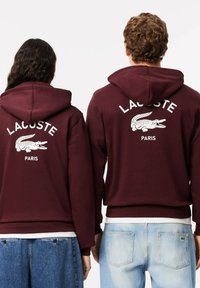 Felpe bordeaux con testo bianco "LACOSTE PARIS" e logo coccodrillo sul retro; indossate con jeans azzurri chiari. Tessuto testurizzato.