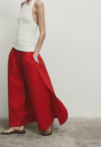 Débardeur en tricot blanc associé à un pantalon large rouge ample avec des fentes latérales. Des sandales noires complètent la tenue.