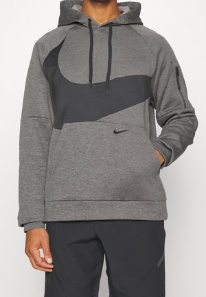 Mand iført en grå og sort Nike-hættetrøje med et stort swoosh-design og kængurulomme, kombineret med sorte bukser.