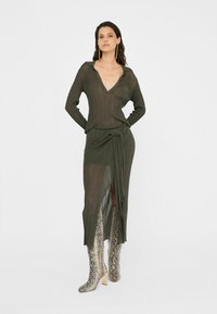 Olivgrünes Strickkleid mit tiefem V-Ausschnitt und langen Ärmeln, das mit einem Wickeldesign und einem seitlichen Schlitz ausgestattet ist, kombiniert mit silbernen Schlangenprint-Stiefeln.