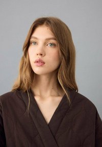 Brauner gesteppter Morgenmantel mit tiefem V-Ausschnitt, weicher Textur und dezentem Steppmuster. Das Modell hat lange Haare und einen neutralen Hintergrund.