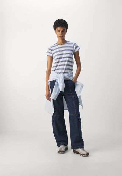 Tricou cu mânecă scurtă, cu dungi alb-negru, asociat cu pantaloni cargo largi din denim închis. Cămașă albastră deschis legată în jurul taliei, adidași neutri.