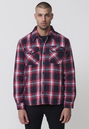 Camisa de franela a cuadros rojos y azul marino con un cierre de botones en la parte delantera, dos bolsillos en el pecho y una etiqueta en la parte superior izquierda. La tela parece texturizada.