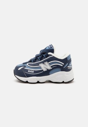 Baskets New Balance bleu marine et blanc avec une semelle épaisse, logo argenté et numéro 1000 sur le côté, montrées de l’extérieur.