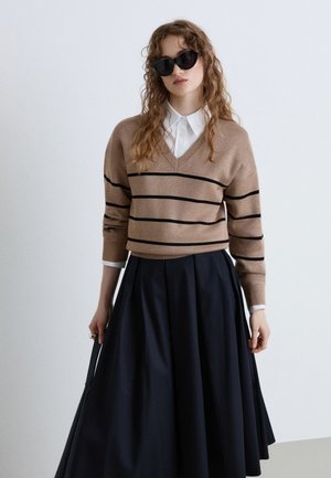 Donna con capelli ondulati che indossa occhiali da sole neri, un maglione a righe beige sopra una camicia bianca e una gonna scura plissettata, che tiene una borsa nera.