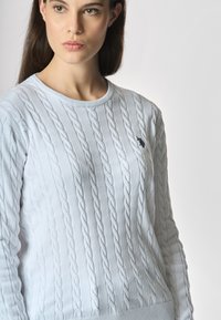 Maglione bianco lavorato a maglia con motivo a treccia, scollo rotondo e sottile dettaglio ricamato di colore scuro sul petto. Texture morbida.