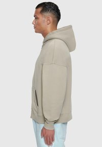 Beige hettegenser med snorhette, kenguru-lomme og ribbestrikkede mansjetter. Glatt stoff, med en avslappet passform og minimale sømdetaljer.