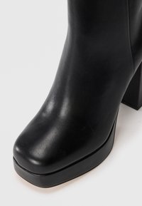Černá kožená kotníková bota se čtveratou špičkou, robustním vysokým podpatkem a mírně zvýšenou podrážkou na bílém pozadí.