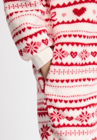 Weißer Fleece-Overall mit roten Herz- und Schneeflockenmuster, ausgestattet mit einem elastischen Handgelenkbund und einer Vordertasche. Weicher, strukturierter Stoff.