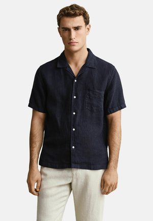 Jeune homme portant une chemise à manches courtes bleu marine à boutons et un pantalon beige clair, debout devant un fond clair uni.