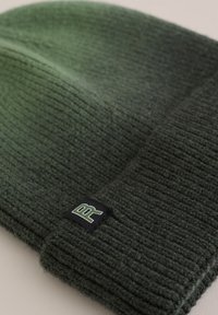 Green knitted beanie with a ribbed texture, featuring a folded cuff and a black patch displaying a white and green logo.