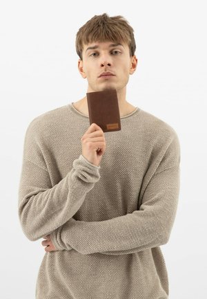 Bruine leren portemonnee met een gestructureerd patroon, vastgehouden door een persoon in een lichtbeige gebreide trui, tegen een eenvoudige witte achtergrond.