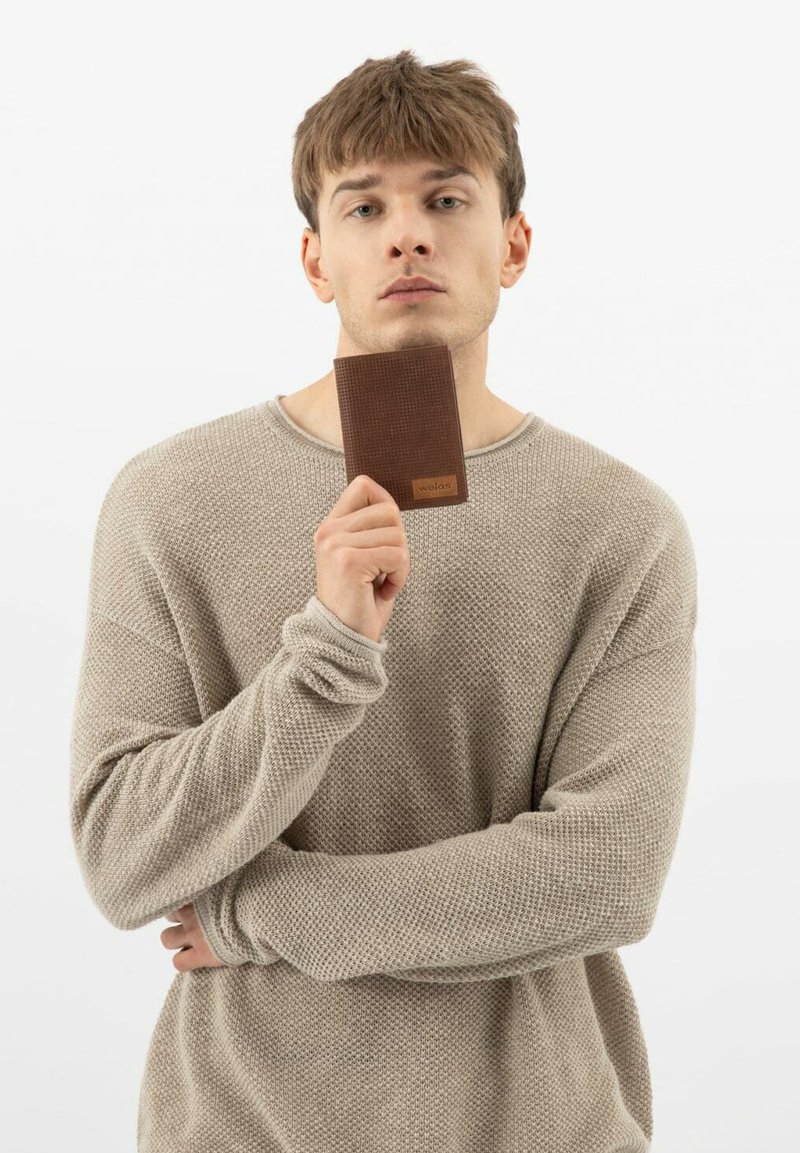 Bruine leren portemonnee met een gestructureerd patroon, vastgehouden door een persoon in een lichtbeige gebreide trui, tegen een eenvoudige witte achtergrond.
