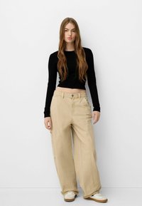 Beige levele cargo dengan potongan lebar yang dilengkapi dengan banyak kantong dan kain yang kokoh, dipadukan dengan atasan hitam lengan panjang yang pas. Sneaker putih melengkapi penampilan.
