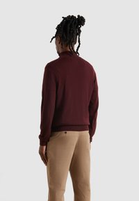 Maglione a maglia color bordeaux con colletto alto, maniche a coste e vita a coste. Abbinato a pantaloni beige con un bottone decorativo sulla tasca.