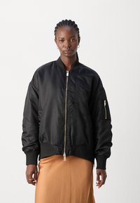 Schwarze Bomberjacke aus glattem Stoff, mit einem Frontreißverschluss, gerippten Bündchen und einer aufgesetzten Tasche auf der Brust, getragen über einem beige Rock.