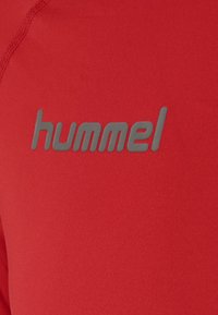 Rød atletisk skjorte med en glat overflade. Logoet "hummel" fremstår i grå og er placeret fremtrædende på forsiden.