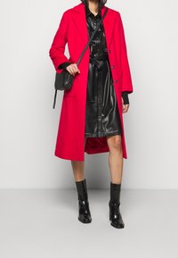 Sytě červený dlouhý vlněný kabát s klopovými límci, zapínáním na jeden knoflík a bočními kapsami, stylizovaný přes černé šaty z umělé kůže a kotníkové boty.