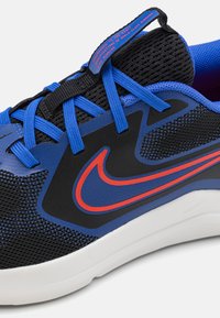 Sportsko med med svart mesh-overdel, blå tekstil-detaljer, og en rød Nike-logo. Har blå snører og såle i hvit gummi.