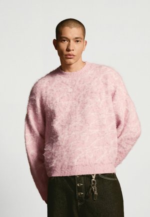 Mand med kort hår iført en blød lyserød sweater og mørke bukser med en nøglekæde hængende fra lommen mod en ensfarvet baggrund.