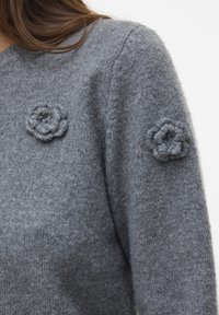 Sanft grauer Strickpullover, der getragen wird und zwei strukturierte Strickblumen-Applikationen aufweist – eine auf der Brust und eine auf dem oberen Ärmel.