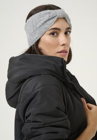 Mujer con cabello oscuro que lleva una diadema de punto gris y una chaqueta acolchada negra, ajustando el cierre de la chaqueta frente a un fondo plano.