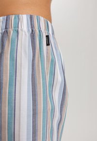 Pantaloni a righe con tonalità di blu, teal, beige e grigio. Vita elasticizzata con una piccola etichetta di marca. Tessuto morbido e texturizzato.