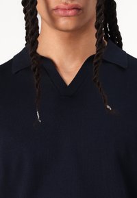 Marinblå stickad polo med V-ringning och korta ärmar, med subtil ribbing vid kragen. Slät textur. Inga synliga mönster.