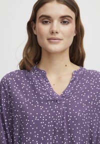 Lila blus med vita prickar, med V-hals och avslappnad passform. Tillverkad av ett mjukt, lätt tyg med slät textur.