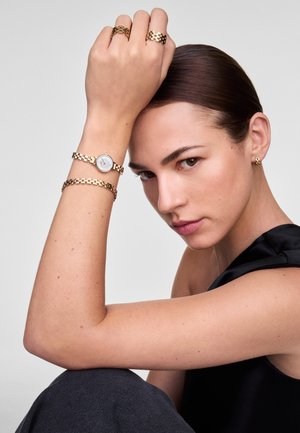 Montre dorée avec un cadran blanc et un bracelet à maillons, accompagnée de bagues dorées assorties et de boucles d'oreilles puces, sur un fond gris.